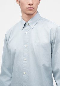 Homme portant une chemise bleu clair à boutons avec un petit logo "BOSS" sur la poitrine gauche, boutons du col fermés, fond uni.