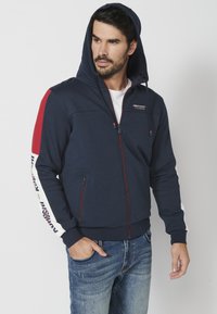 Jūrinės mėlynos zip-up sportinės striukės su raudonais akcentais ant pečių ir rankovių, su priekinėmis kišenėmis, kapisonu su raišteliais ir logotipu ant krūtinės.