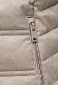 Šviesiai pilkos spalvos paminkštintas švarkas su subtiliu raštu ir užtrauktuku su prekės ženklo etikete. Matomas tekstūruotas ir siūtas paviršiaus detalė.