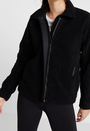 Frau trägt eine schwarze Sherpa-Fleecejacke mit Reißverschluss über einem weißen Shirt und schwarzen Leggings, steht vor einem einfarbigen Hintergrund.