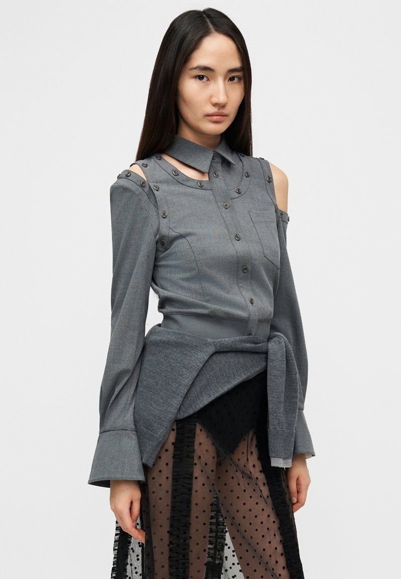 Grå struktureret skjorte med knapdetaljer, lange ærmer og kold skulder-design, kombineret med en gennemsigtig sort nederdel med prikker.