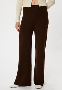 Bruine, wijde broek van een textuurstof met een hoge taille. Gepaarde met lichte schoenen en een ribbeltrui in crème.