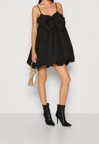 Svart ärmlös minikjol med en strukturerad yta och ett stort båtdetalj fram. Matchad med svarta ankelstövlar och en guldfärgad handväska.