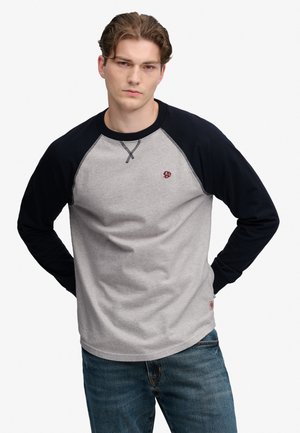 Jonge man met kort gestyled haar, draagt een grijs-zwart shirt met lange mouwen en blauwe jeans, staat met de handen achter zijn rug.