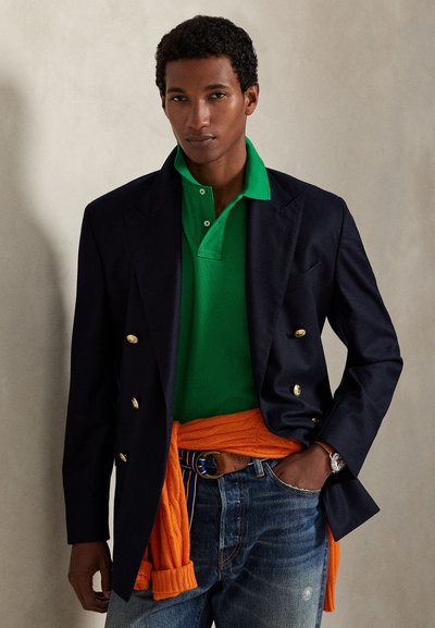 Μπλε ναυτικό ζακέτα διπλής σειράς κουμπιών πάνω από πράσινο πόλο, πορτοκαλί πλεκτό πουλόβερ γύρω από τη μέση, μπλε τζιν και ρολόι, σε ουδέτερο φόντο.