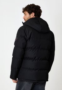 Doudoune noire avec capuche, ornée d'un design matelassé, de poignets élastiques et d'un écusson logo sur l'épaule. Texturée, chaude et isolante.