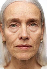 Close-upportret van een oudere vrouw met wit haar, groene ogen en lichte huid. Ze heeft zichtbare rimpels en draagt gouden oorringen.