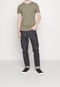 Muž v olivově zeleném tričku s krátkým rukávem, tmavých ohrnutých džínách a černých teniskách stojící s jednou rukou v kapse na bílém pozadí.