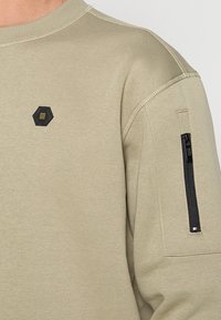 Olivengroene sweatshirt met een gestructureerde afwerking, voorzien van een zwart zeshoekig patch en een met rits afsluitbaar zakje op de linker mouw voor functionaliteit.