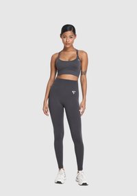 Žena stojí čelom vpred, oblečená v tmavosivom športovom podprsenke a legínach s bielymi teniskami na svetlom pozadí.