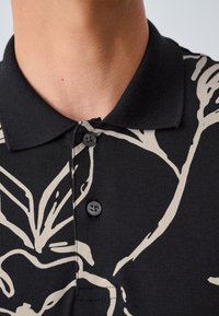 Juodas polo marškinėlis su smėlio spalvos abstrakčiu raštu, su trijų sagų užsegimu ir tekstūruotu apykakle. Audinys atrodo minkštas ir lengvas.