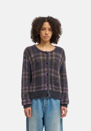 Personne aux cheveux bouclés portant un cardigan gris à carreaux duveteux, avec des lignes violettes et jaunes, et un jean bleu clair, debout sur un fond uni.