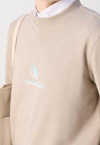 Persona che indossa un maglione beige dei Calvin Klein Jeans sopra una camicia bianca, con una tracolla di una borsa tote beige sulla spalla.