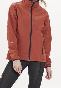 Röd softshell-jacka med hög krage, marinblå dragkedjor och elastiska ärmslut. Inkluderar sidofickor och en logotyp på fållen.