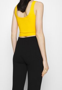 Žlutý žebrovaný crop top s širokými ramínky, spárovaný s černými žebrovanými kalhotami s vysokým pasem. Hladká textura, přiléhavý střih.