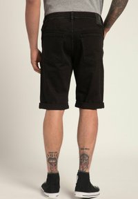 Shorts in denim nero con orli arrotolati, dotati di due tasche posteriori. Indossati con sneakers alte nere. Tatuaggi visibili sulle gambe inferiori.