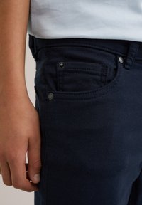 Tamsiai mėlyni džinsai su siauru siluetu, priekinėmis kišenėmis su metaliniais nugariniais ir kontrastinėmis siūlėmis.