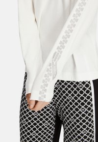 Hvid langærmet top med et tekstureret mønster langs ærmet, kombineret med sorte bukser med et gentaget hvidt cirkulært grafisk design.