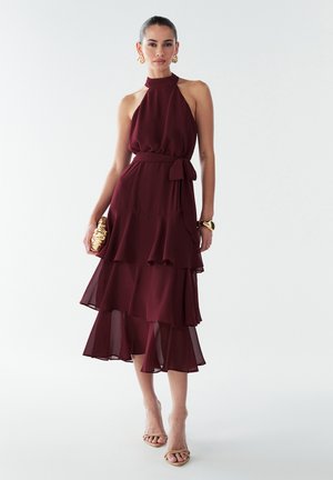 Robe bordeaux à col halter, avec un ourlet à volants superposés, une ceinture à la taille et un tissu fluide. Associée à des accessoires dorés et des talons nus à lanières.