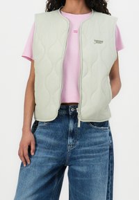 Vaaleanvihreä tikattu liivi vetoketjulla, jossa hienovarainen logo. Päällä vaaleanpunainen t-paita ja yhdistetty sinisiin farkkuihin.