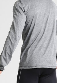Camiseta deportiva de manga larga en gris, con una textura suave, dobladillo curvado y costuras reforzadas. Sin logotipos ni patrones visibles.