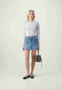 Top scurt, cu maneci lungi, gri deschis; fustă mini din denim cu margine desfăcută; încălțăminte plată negre cu studs; geantă gri cu model. Fundalul este neutru.