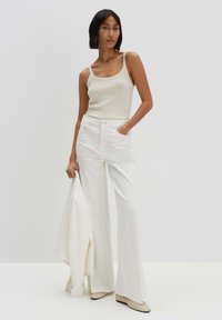 White wide-leg pants made of smooth fabric, paired with a fitted beige tank top. Model holds a white jacket, wearing light beige shoes.