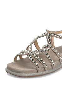Sandalia de ante beige con tiras entrecruzadas adornadas con tachuelas plateadas, una plantilla texturizada y una base plateada con brillo.