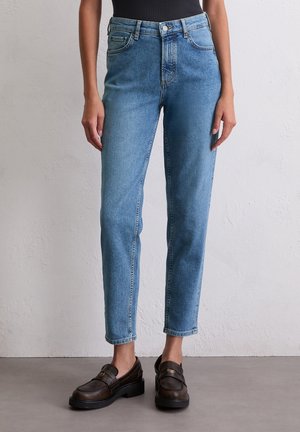 Woman wearing high-waist blue denim jeans with front pockets and button closure, paired with black loafers and cropped black top.