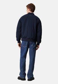 Giacca bomber blu navy con orlo e polsini aderenti, caratterizzata da una texture liscia e un design semplice. Abbinata a jeans azzurri chiari.