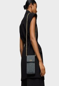 Bolso cruzado de textura azul oscuro con una cadena dorada y solapa frontal, que presenta el nombre de la marca en relieve "TOUS". Diseño simple y rectangular.