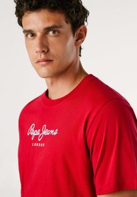 Rdeča majica s kratkimi rokavi iz bombaža, z belim napisom "Pepe Jeans London" čez prsa. Gladka tekstura in okrogla izrezana ovratnika.