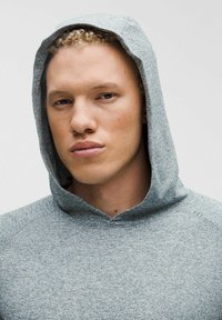 Sivá mikina s textúrovanou tkaninou, kapucňou a vysokým golierom. Dizajn má jednoduchý, moderný štýl bez viditeľných log alebo vzorov.