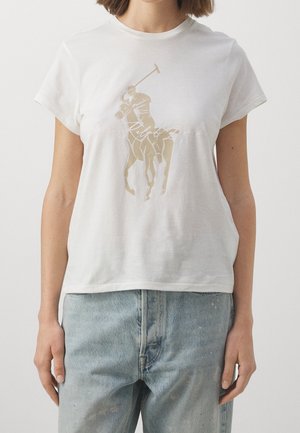 Kvinde iført hvid t-shirt med korte ærmer med grafisk motiv af beige polo-spiller og lyseblå højtaljede jeans mod ensfarvet baggrund.