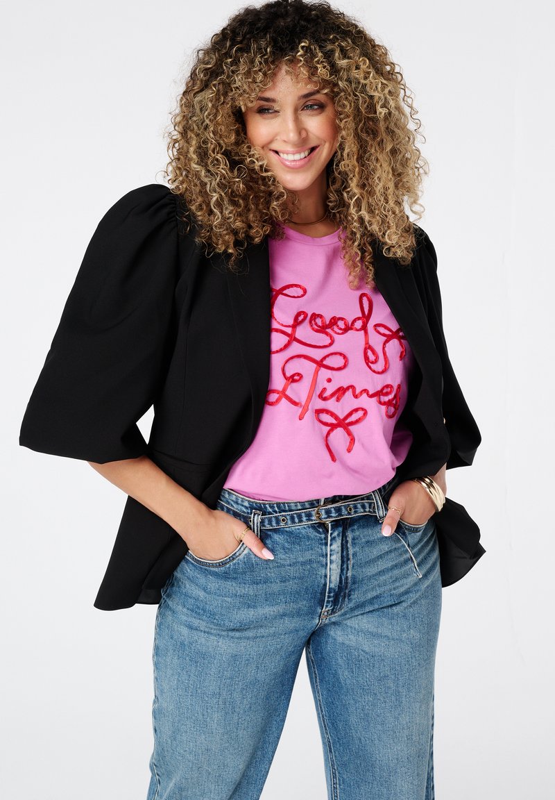 Zwarte blazer met pofmouwen over een roze T-shirt met rode letters "Good Times," gecombineerd met hooggeknipte blauwe jeans.