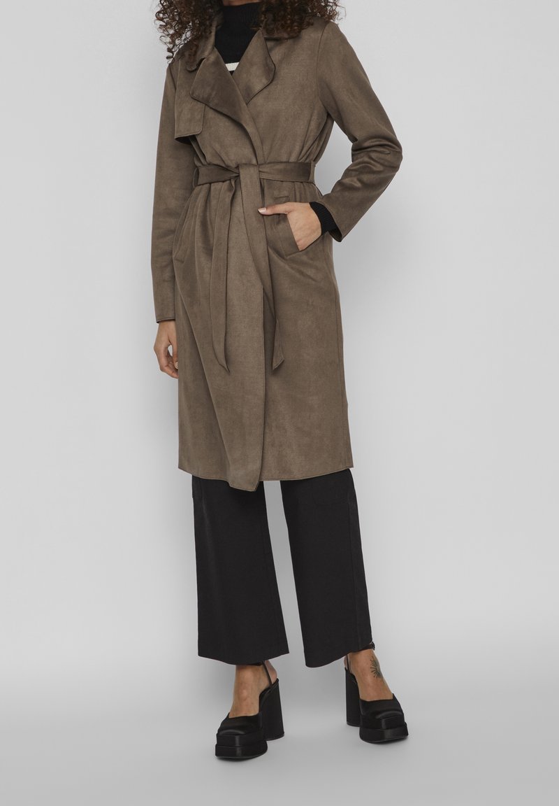 Trench-coat marron clair en daim avec une ceinture à la taille, de grands revers et des poches latérales, porté avec un pantalon noir à jambes larges et des chaussures à plateforme.