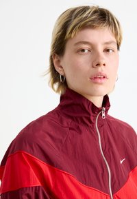 Rød og burgunder vindjakke med høy krage, glidelås foran og sidepaneler. Har en hvit Nike-logo på brystet.