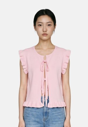 Donna che indossa un top senza maniche rosa chiaro con bordi arricciati e lacci frontali, abbinato a jeans di denim blu su uno sfondo bianco semplice.