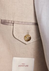 Veste en lin beige avec une poche poitrine ornée d'un bouton glossy de couleur marron clair. L'étiquette indique "NEXT SIGNATURE". Texture lisse avec un tissage subtil.