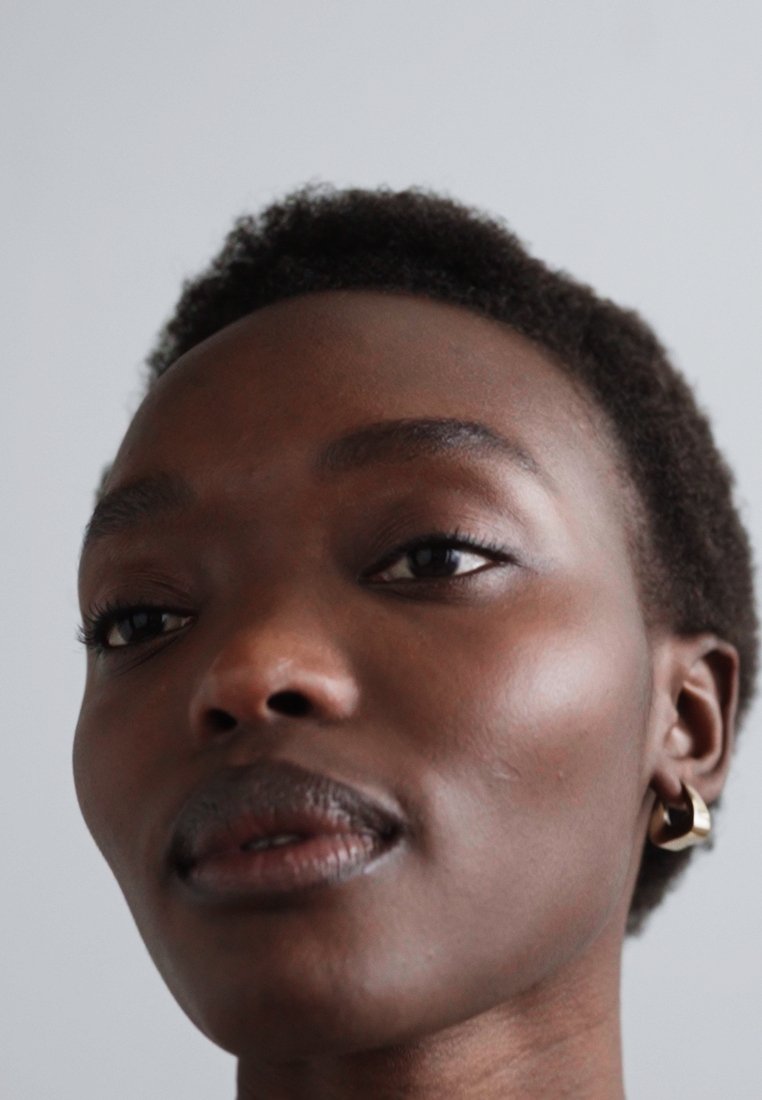 Gros plan d'une personne aux cheveux courts et bouclés, de couleur foncée, portant une boucle d'oreille en or, avec une peau lisse et un maquillage naturel, sur un fond clair.