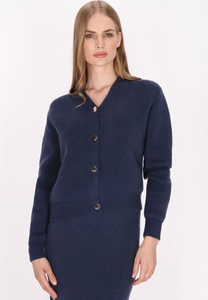 Tmavě modrý pletený cardigan s véčkovým výstřihem, dlouhými rukávy a zavíráním na pět zlatých knoflíků. Obsahuje žebrované manžety a spodní lem pro přiléhavý vzhled.