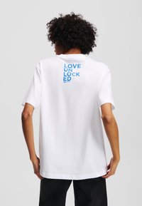 Bílé bavlněné tričko s nápisem "LOVE UNLOCKED" v modré na zadní straně. Klasický střih s kulatým výstřihem, krátké rukávy a uvolněný střih.