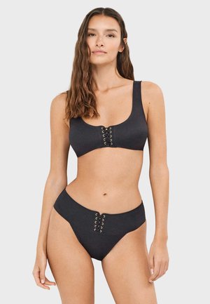 Femme aux longs cheveux bruns portant un maillot de bain deux-pièces noir avec des détails à lacets en haut et en bas sur un fond clair uni.