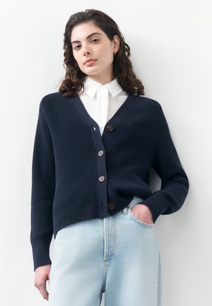 Junge Frau mit lockigem dunklem Haar, die ein weißes Hemd mit Kragen, eine marineblaue Strickjacke mit Knöpfen und hellblaue Jeans trägt und eine Hand in der Tasche hat.