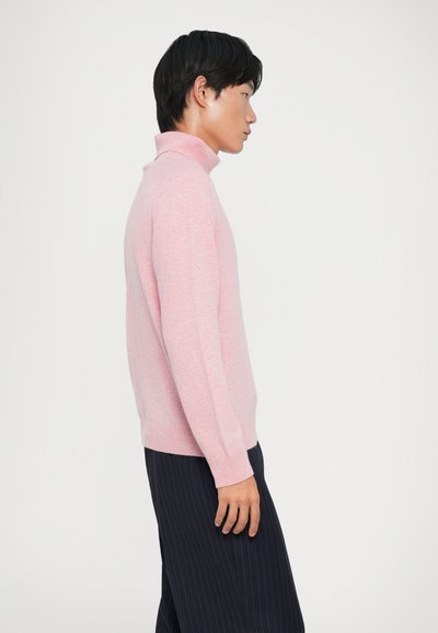 Jasnoróżowy sweter typu golf w miękkim dzianinowym materiale, dopasowany krój, ściągacze na rękawach, w zestawieniu z ciemnymi spodniami w prążki. Widok z boku.