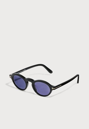 Lunettes de soleil noires à monture ronde avec verres bleu foncé et branches fines posées sur une surface claire.