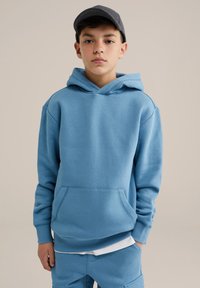 Felpa blu con cappuccio realizzata in tessuto morbido, dotata di tasca frontale e polsini a costine. Indossata con pantaloni jogger blu coordinati.