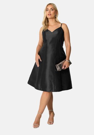 Robe en satin noir avec un décolleté en V, des bretelles fines et une jupe évasée. Le mannequin tient une pochette métallique texturée et porte des talons argentés à lanières.