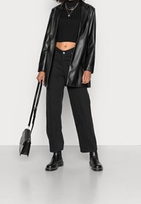 Blazer in pelle nera sopra un top corto nero a coste, abbinato a pantaloni neri a gamba larga e stivaletti neri alla caviglia. Gli accessori includono collane a strati.