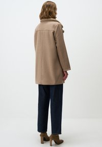 Cappotto di lana beige con colletto alto, maniche lunghe e tasche laterali, abbinato a jeans a gamba larga blu scuro e stivaletti con tacco color crema.